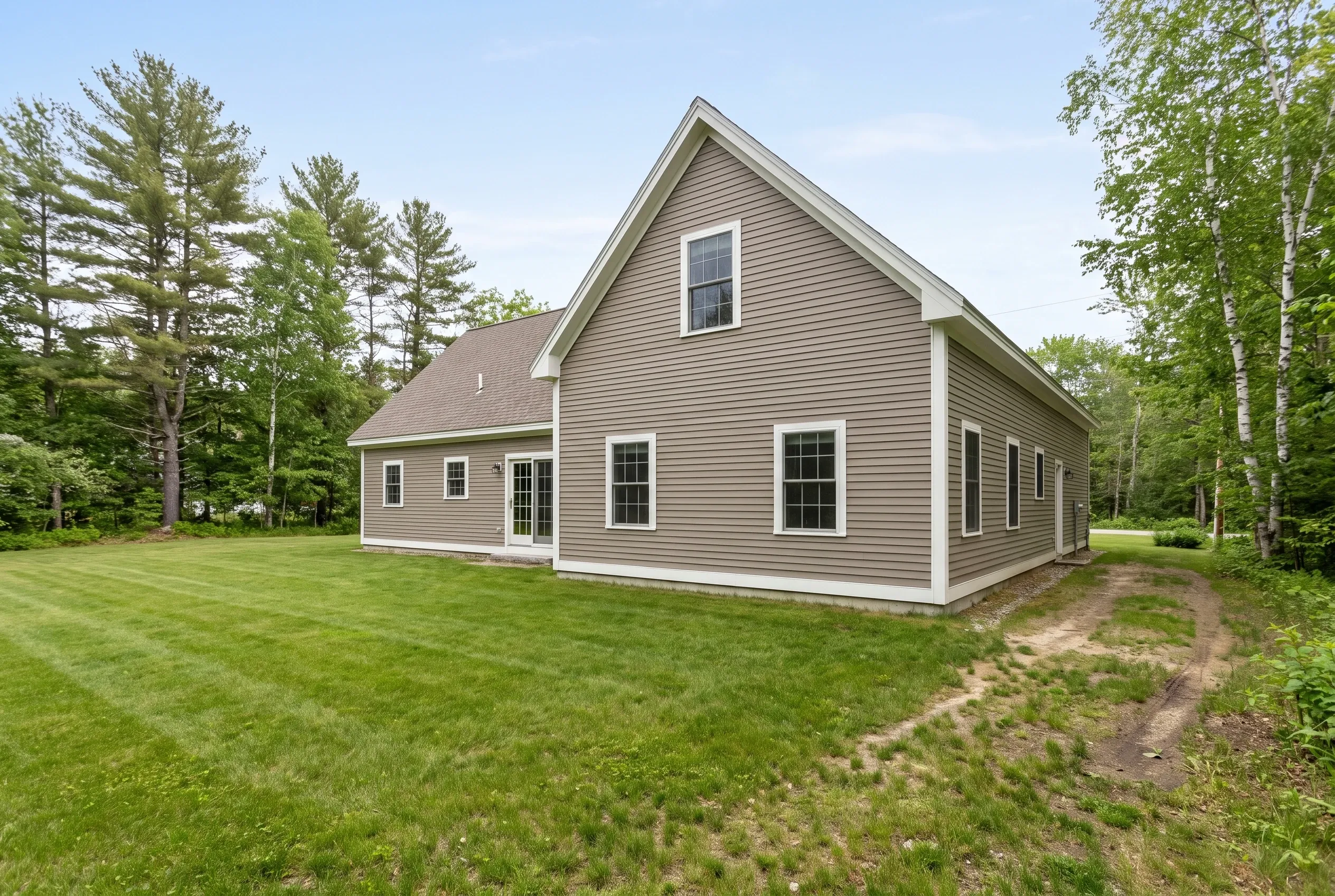 Taupe-sided house with white trim behind a mowed lawn with worn dirt tire tracks, surrounded by pine and birch trees