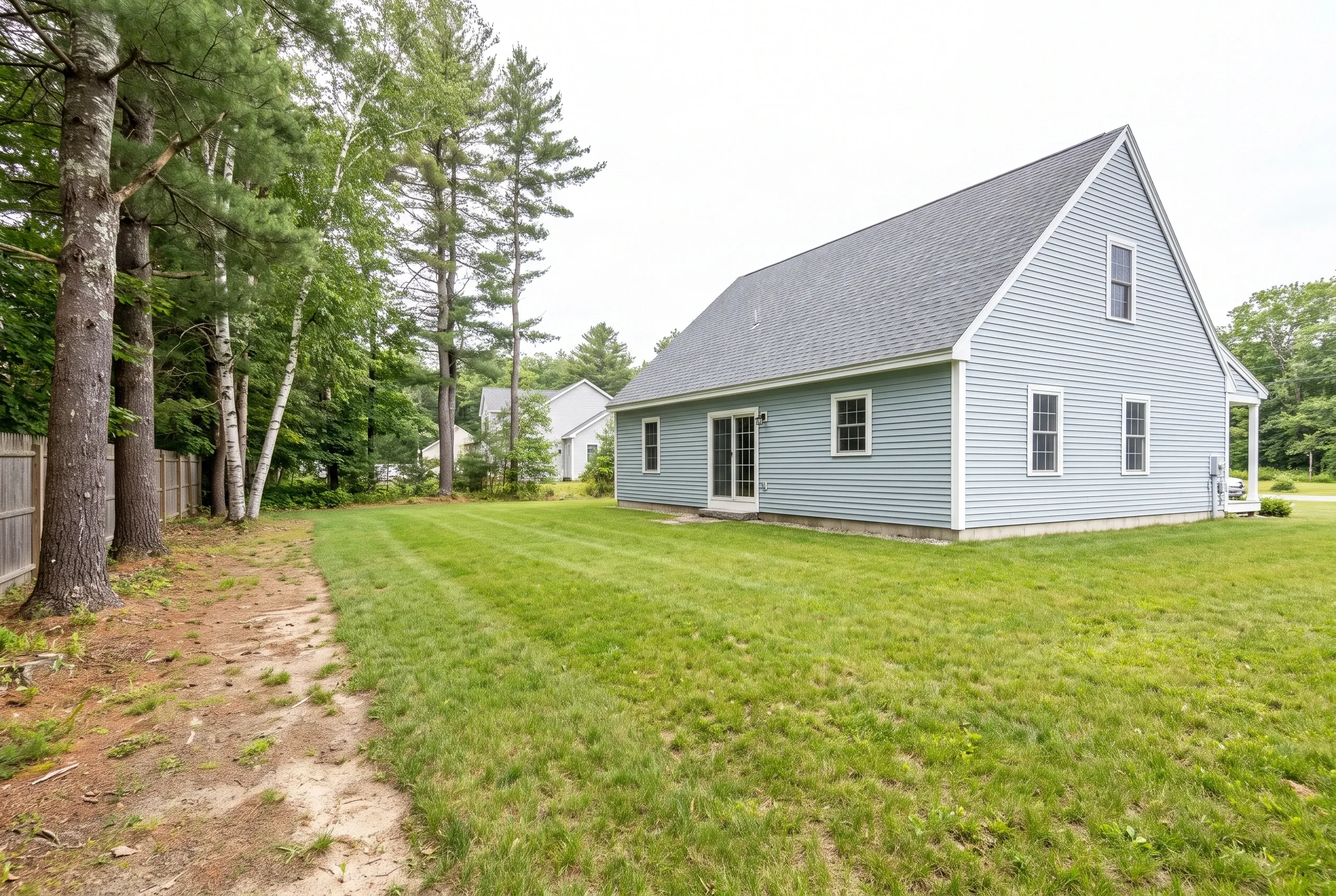 Open backyard lawn beside a light blue gable house, with a worn dirt path and tall pine and birch trees