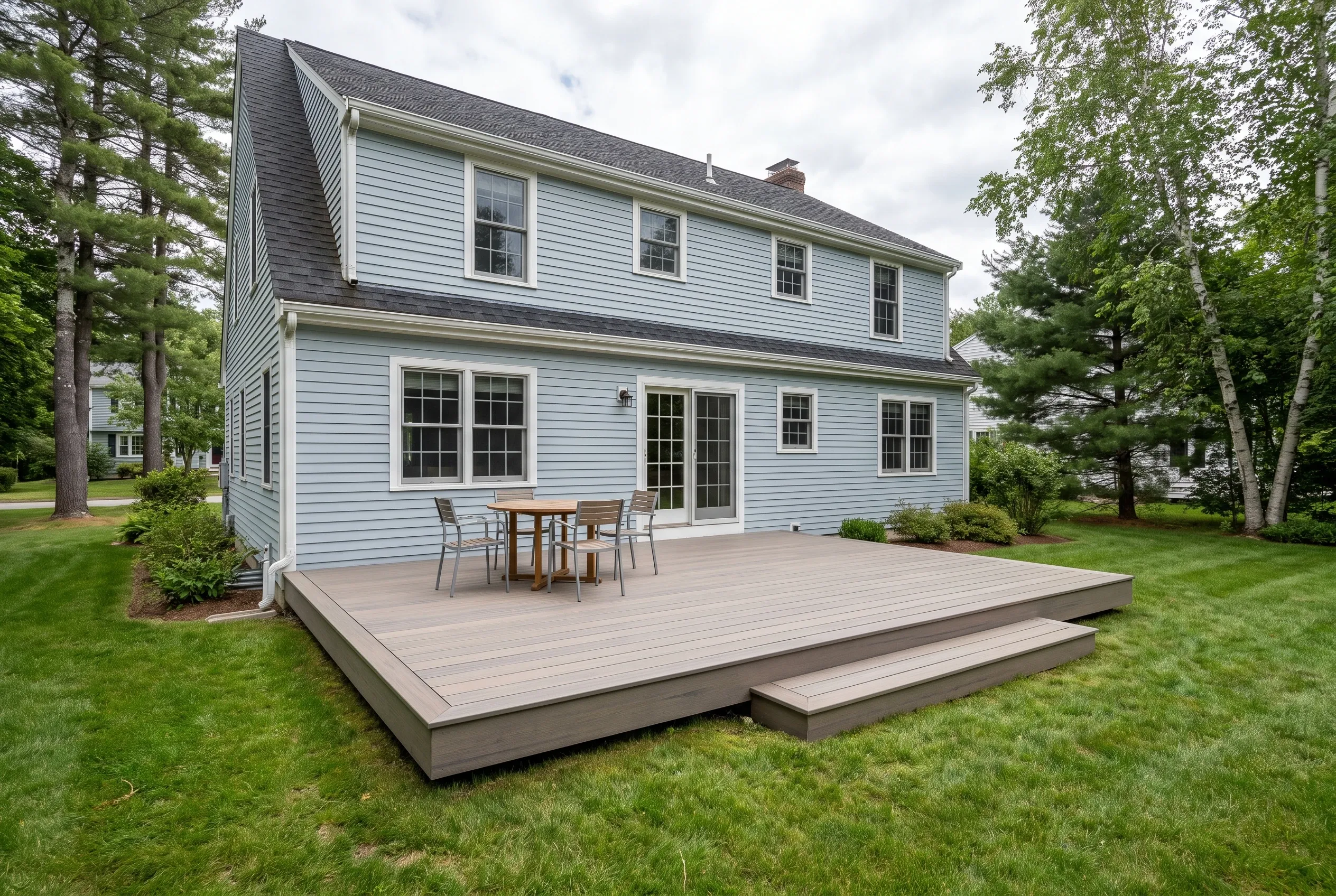 Deck builders in Eliot, ME: low gray deck with a step, table and chairs behind a light blue two-story house