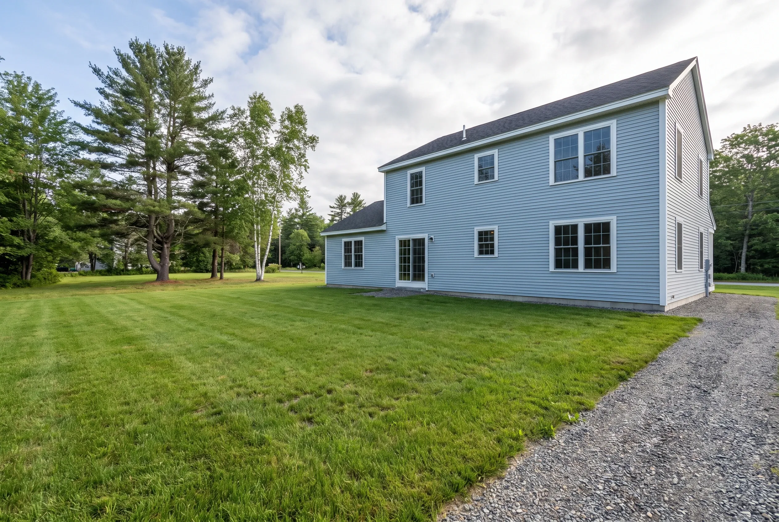 Rear view of a light blue two-story house with an open lawn, gravel driveway, and tall pine and birch trees