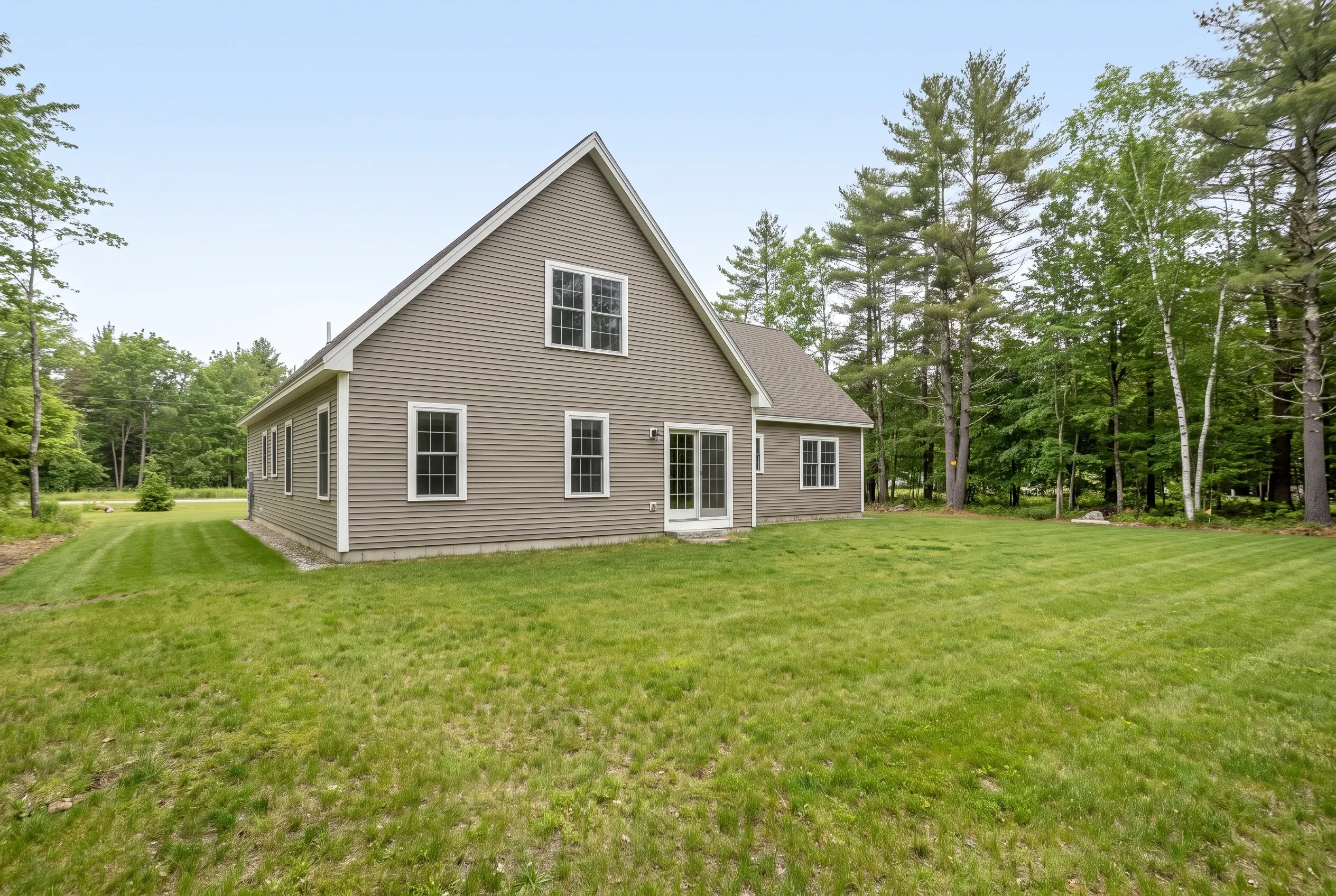 Taupe-sided house with a gabled roof and an open mowed backyard lawn bordered by pine and birch trees, no deck present