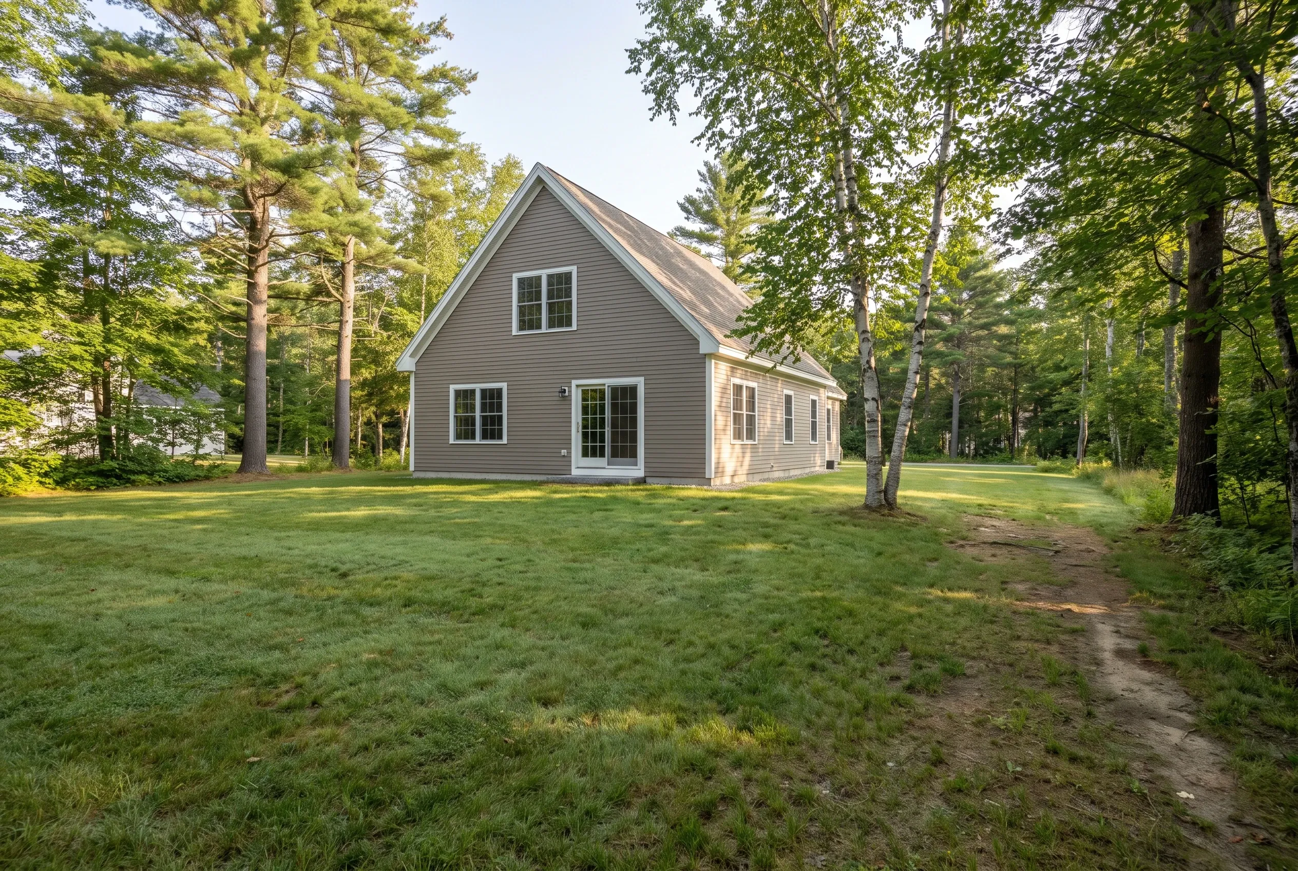 Open grass backyard with a worn footpath leading toward a tan two-story house surrounded by pine and birch trees