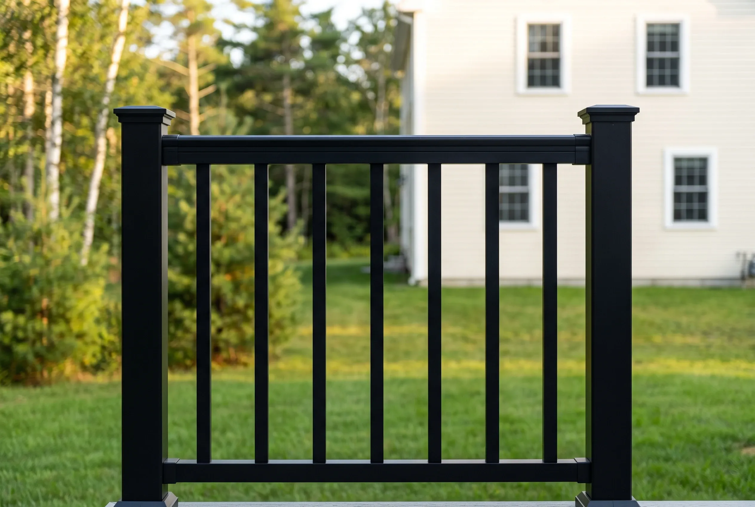 Close view of a black railing section with square posts and vertical balusters, with lawn, trees and a white house behind