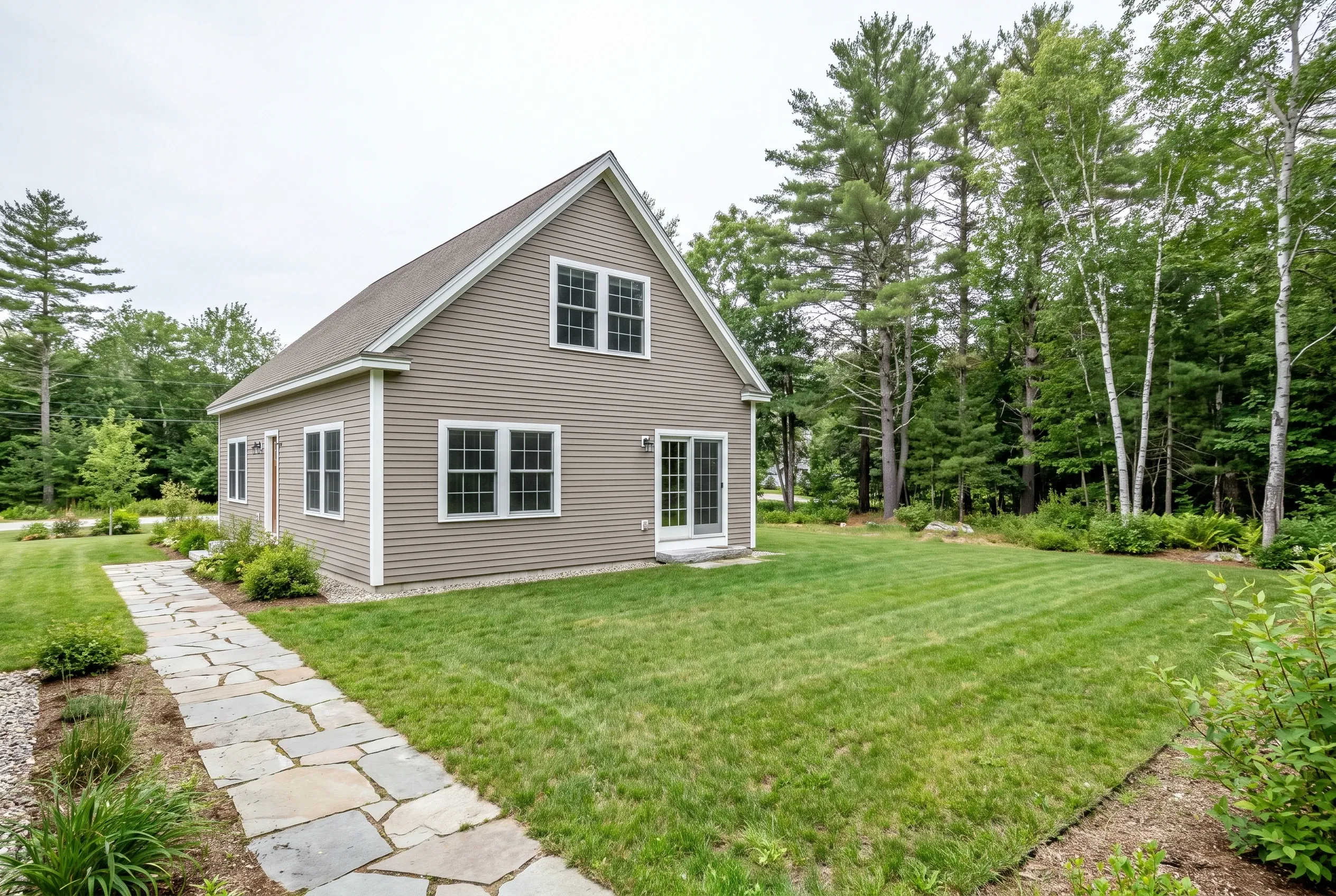 Backyard of a gray-sided house with a flagstone path, open lawn, and surrounding pine and birch trees before any deck