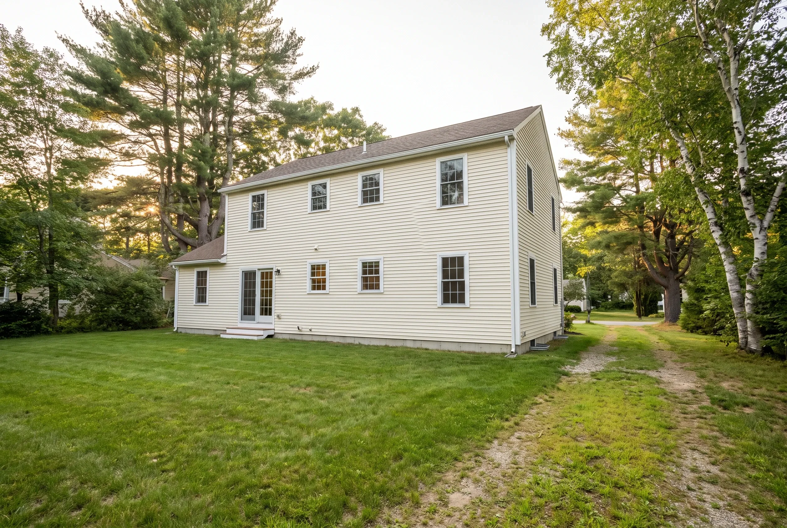 Open grassy backyard behind a two-story cream clapboard house with trees and a dirt path, no deck present