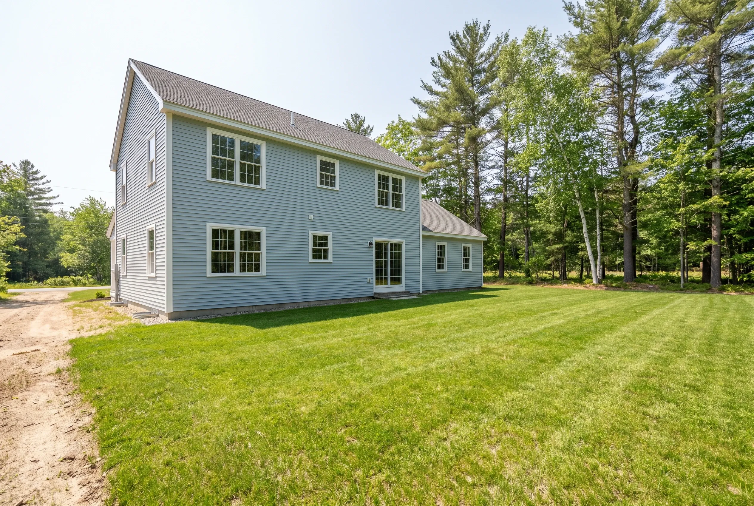Two-story light blue house with white trim beside a wide mowed lawn, dirt driveway, and tall pine and birch trees