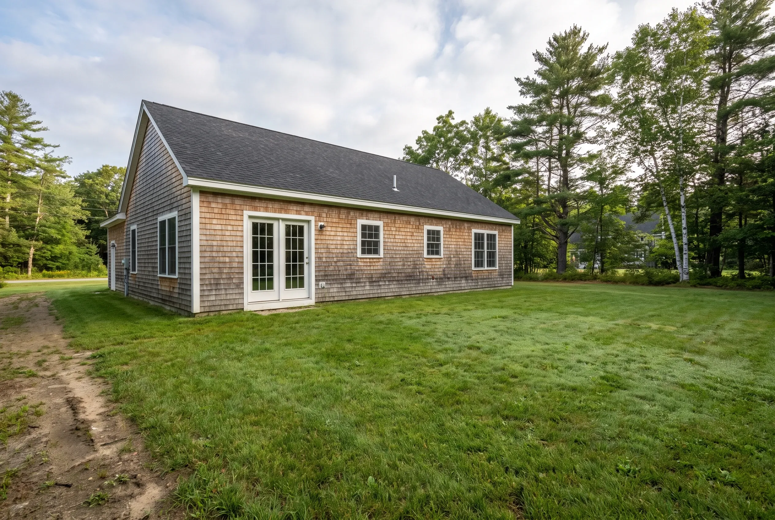 Single-story shingled house with French doors facing an open lawn, a worn dirt path, and pine and birch trees