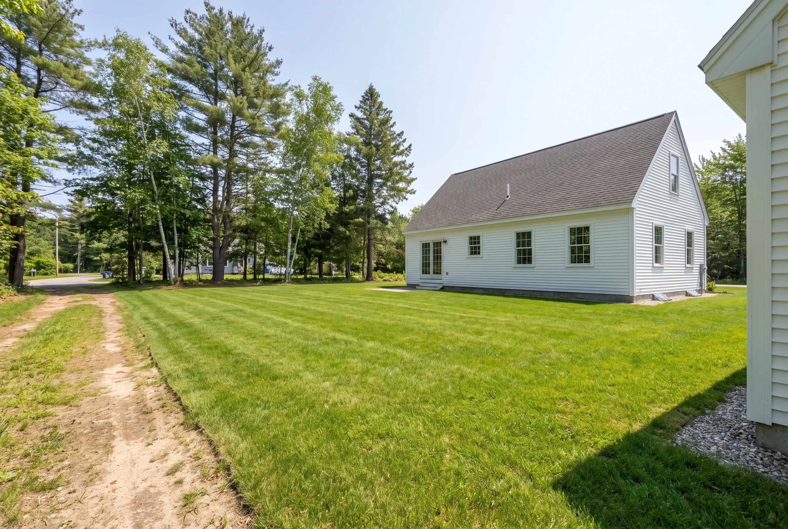 Open backyard lawn with a dirt path, white gabled house, and tall pine trees; edge of another white house wall at right