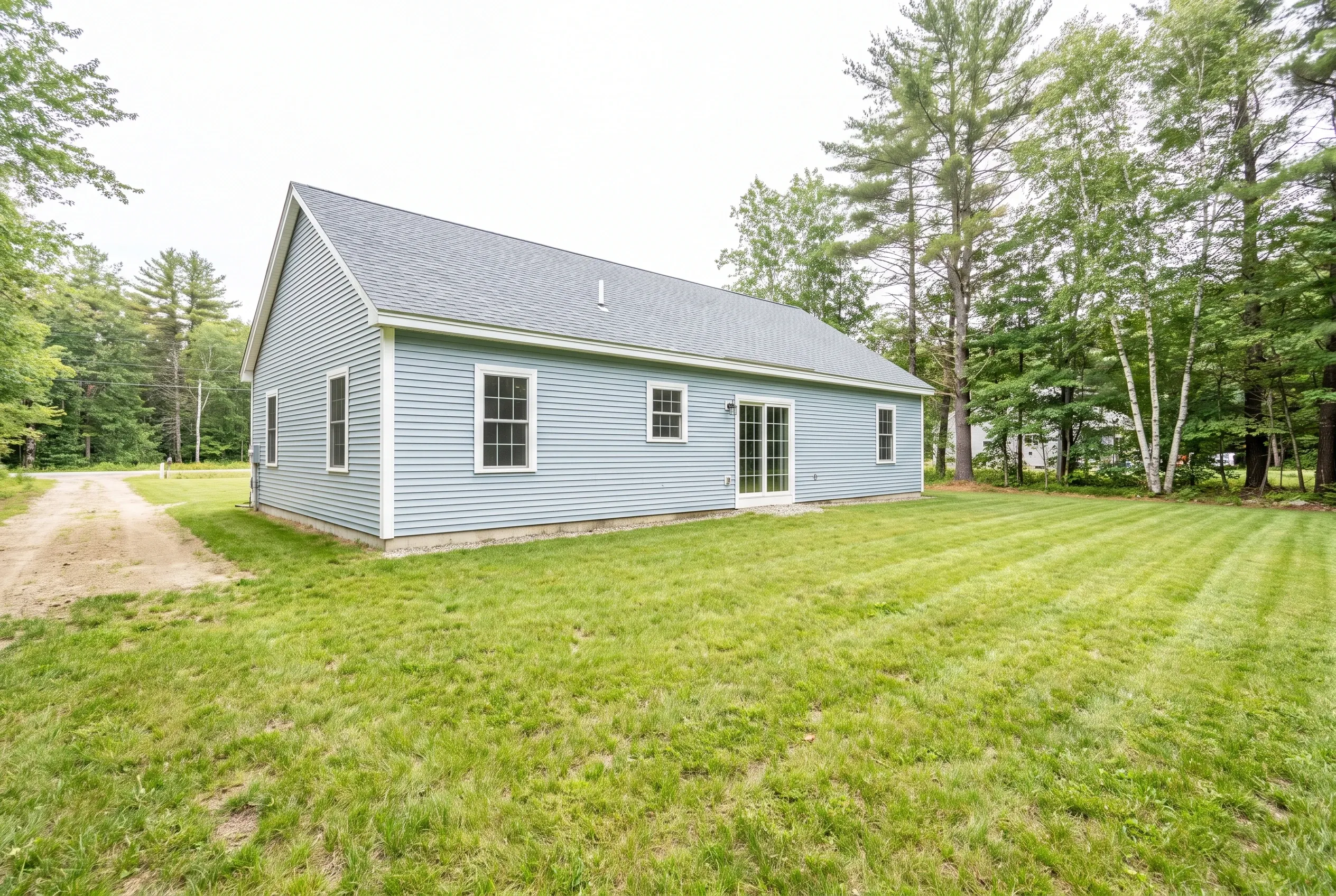 Light blue single-story house with rear patio door, open mowed lawn, gravel driveway and surrounding pine and birch trees