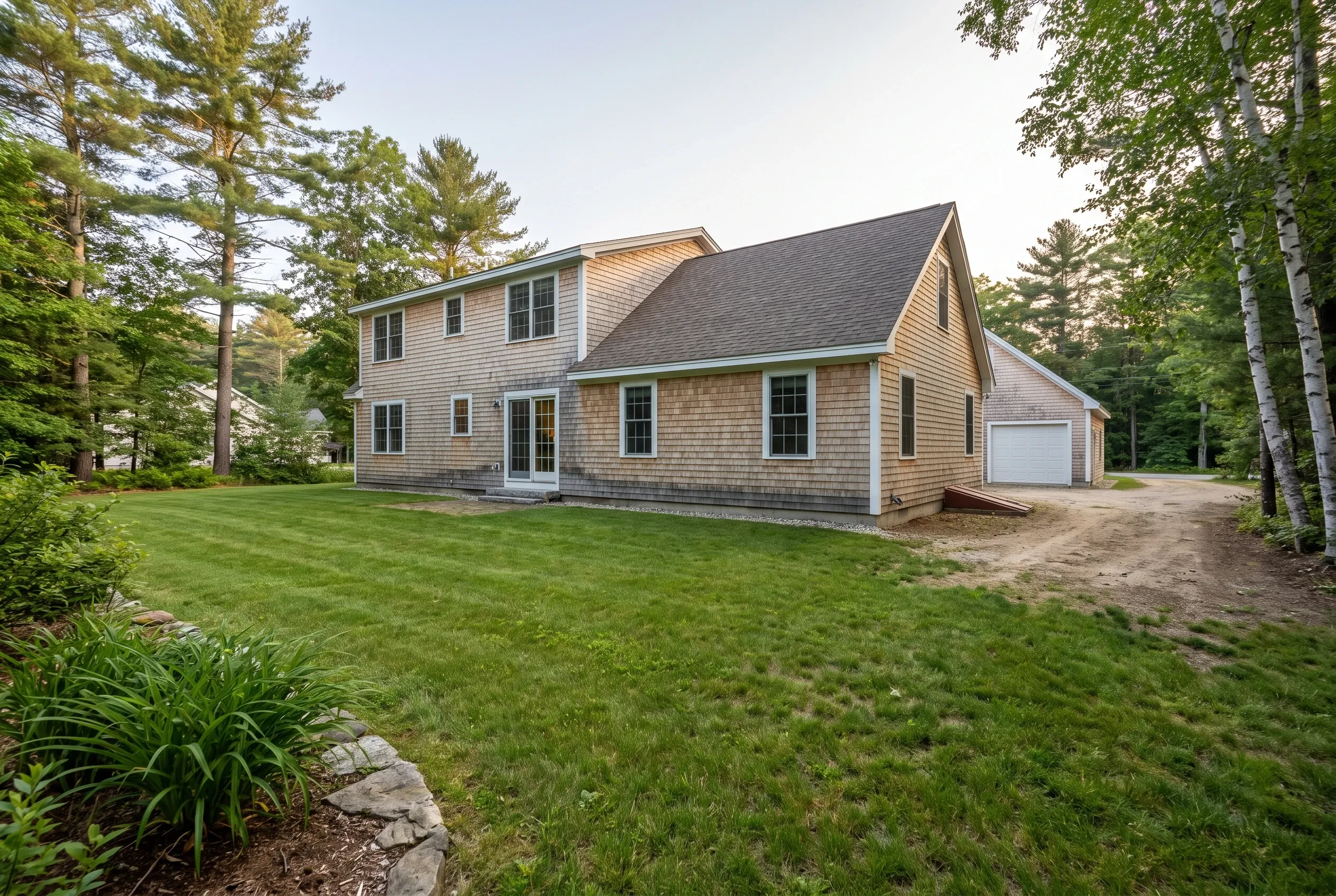 Backyard of a shingled two-story house with detached garage, dirt driveway area, open lawn, and surrounding pine and birch trees